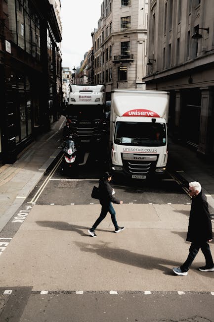 A narrow city street scene during daytime featuring a white removal van with the logo and website of Removal Van Earls Court parked partially on the pavement and partially over traffic lanes. The van is positioned close to a building on the right side, with its rear doors likely open for loading or unloading furniture and boxed items. A scooter is parked beside the van, and two pedestrians are walking across a zebra crossing in the foreground, one with a backpack and the other in a dark coat. The street is lined with multi-storey buildings with a mix of modern and older architectural styles, and the environment appears bright with natural daylight. This scene illustrates the logistics involved in home relocation, furniture transport, and packing during a removal process, highlighting the use of a removal company's vehicle for efficient moving services in the Earls Court area.
