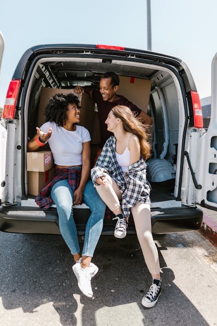 Two young women sitting on the edge of an open moving van parked outdoors, engaging in conversation and smiling, with packed cardboard boxes and soft furnishings visible inside the vehicle. The woman on the left has curly hair and is wearing a light blue t-shirt and jeans, while the woman on the right has long hair and is dressed in a white top, light-colored leggings, and sneakers, with a plaid shirt tied around her waist. The van, part of the services provided by Removal Van Earls Court, is situated near a curb on a sunny day, illustrating the home relocation process involving furniture transport, packing, and loading activities typical of professional removals teams.