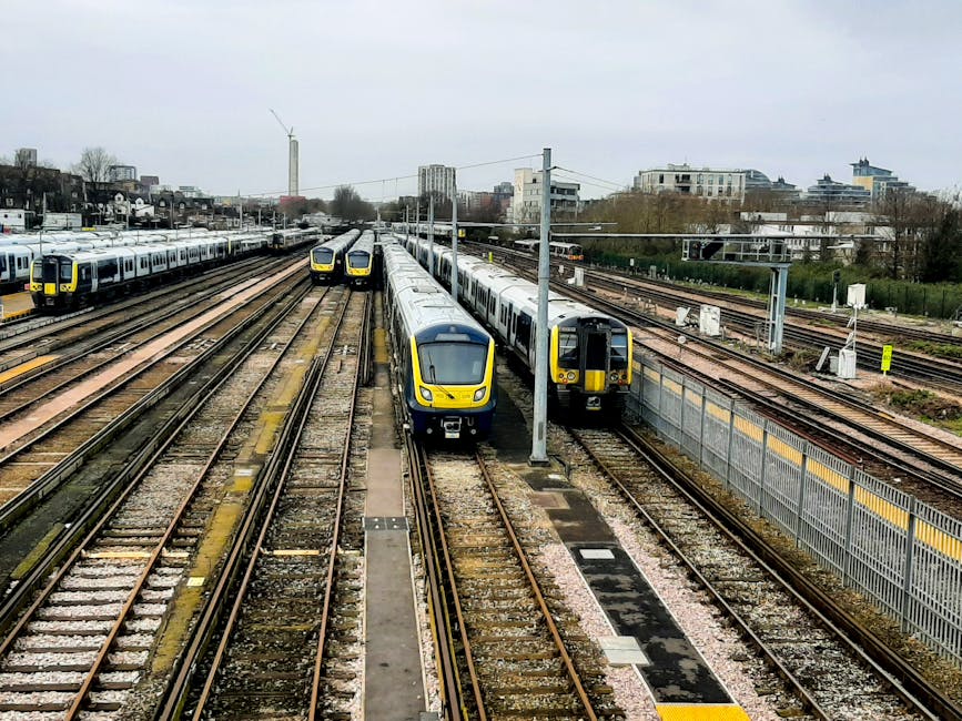 A view of multiple modern passenger trains with yellow and black accents parked on railway tracks near Earls Court station in London. The trains are aligned in parallel lines, with some facing towards the camera and others pointing away, situated on a busy railway yard. Overhead electrical wires and poles run above the tracks, which are surrounded by a mix of urban infrastructure including fencing, small buildings, and residential or commercial structures in the background. The sky is overcast, and the scene appears to be during daytime with diffuse natural light. This setting reflects the context of home relocation and moving logistics, as the railway yard is associated with transporting goods or furniture during a house move. Occasionally, Removal Van Earls Court may utilize such transport infrastructure for efficient furniture transport and logistics management during house removals near Earls Court station.