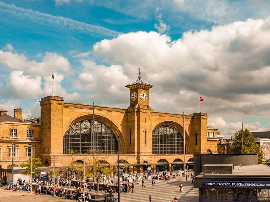 Image of King's Cross railway station in London, showing a large brick building with arched windows and a central clock tower with a weather vane on top. The station is busy with people walking on the